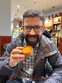 A smiling man with a short beard and glasses sitting in a café with a small pie in his hand.