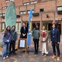 Group of Student Ambassadors smiling and standing by a Robinson crest flag and a sign which reads 'Offer Holder Tours Meeting Point' 