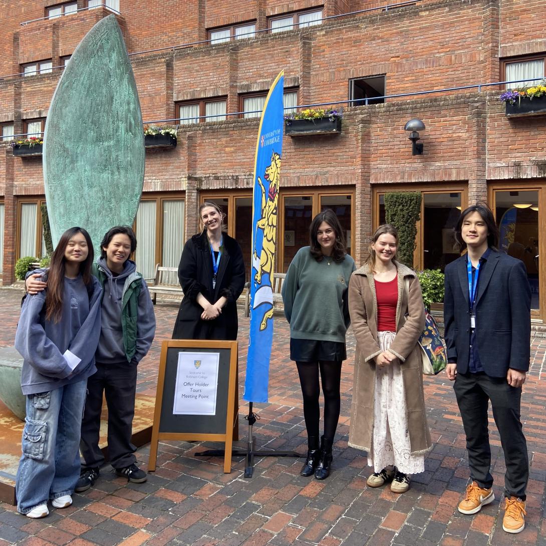 Group of Student Ambassadors smiling and standing by a Robinson crest flag and a sign which reads 'Offer Holder Tours Meeting Point' 